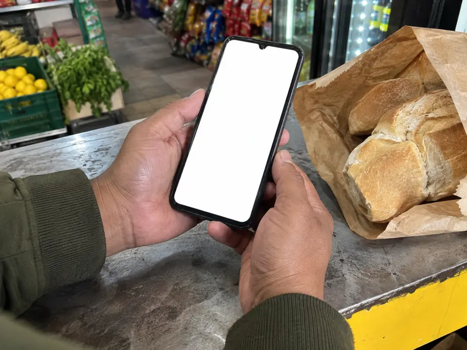 Manos tomando un celular sobre el mostrador de un almacén, junto a una bolsa de pan